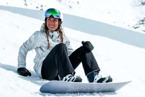 Uśmiechnięta snowboardzistka w kasku i goglach siedzi na śniegu obok deski. Jej pokrowiec na deskę snowboardową jest gotowy do drogi.