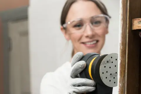 Mujer con gafas de seguridad usa lijadora orbital para restaurar madera, una forma eficiente de como lijar madera sin lija.