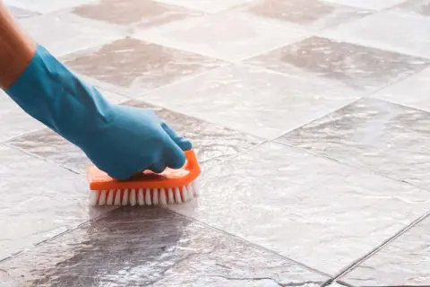 Mano con guante azul limpiando suelo de baldosas con cepillo naranja. Solución para con que fregar el suelo si no tengo fregasuelos.