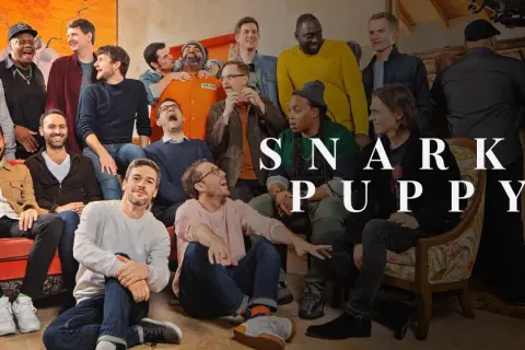 Grupa ludzi, w tym mężczyzna w pomarańczowym kombinezonie, pozuje do zdjęcia z napisem "SNARKY PUPPY".