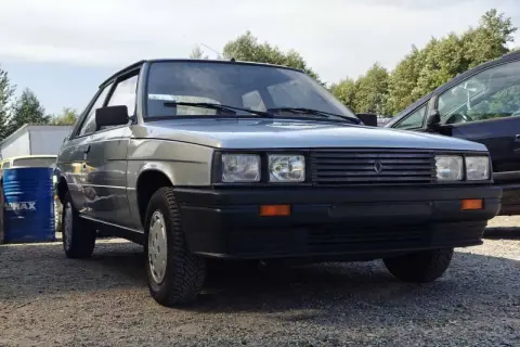 Srebrny Renault 11 z lat 80. stoi na żwirowym podjeździe, obok niebieskiego beczki i innego samochodu.