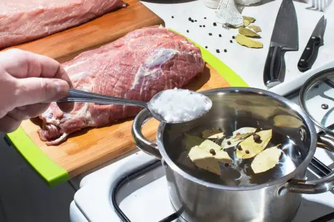 Przygotowanie solanki do mięsa: ręka wsypuje sól do garnka z wodą, liśćmi laurowymi i pieprzem. Obok surowe mięso na desce.