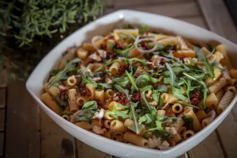 Ein köstlicher nudelsalat mit getrockneten tomaten, Rucola und Mozzarella in einer weißen Schüssel.