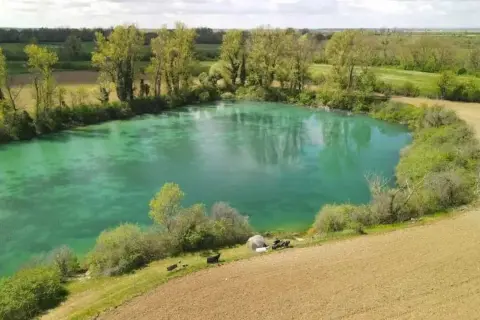 Idyllischer Paylake in Frankreich mit türkisfarbenem Wasser, umgeben von grüner Vegetation und Feldern. Angler haben ihr Lager aufgeschlagen.