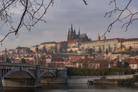 Praga nietypowe atrakcje: widok na Hradczany i Most Karola przez Wełtawę, z gałęziami drzew w pierwszym planie.