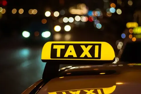 Żółty znak TAXI na dachu samochodu nocą. W tle rozmyte światła miasta, sugerujące licencję taxi w ruchu.