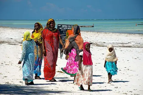 Kobiety i dzieci w kolorowych strojach spacerują po plaży Zanzibaru. Przed wyjazdem warto pomyśleć o zanzibar szczepienia.