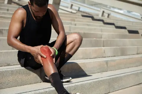 Sportler mit schmerzhaft gerötetem Knie auf Treppen. Er hofft, seine Gelenkschmiere aufbauen zu können, um bald wieder fit zu sein.