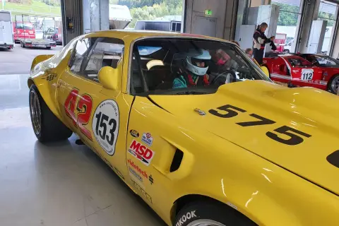 Gul Firebird 69 med nummer 153 och 575 redo för race. En förare i hjälm sitter bakom ratten.