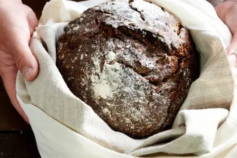 Ręce trzymają bochenek chleba żytniego, który sugeruje, jaka mąka jest najzdrowsza. Posypany mąką, zawinięty w lniany worek.