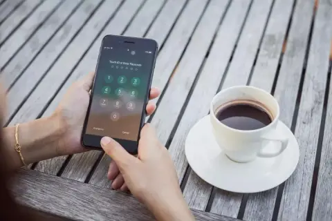 Zablokowany iPhone z klawiaturą numeryczną i ikoną Touch ID. Obok filiżanka kawy na drewnianym stole.