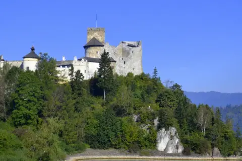 Zamek w Niedzicy, majestatyczna twierdza na wzgórzu, otoczona zielenią, z widokiem na jezioro i góry.