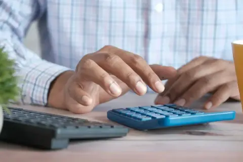 Hände tippen auf einem blauen Taschenrechner, während eine gelbe Tasse und eine Tastatur daneben stehen. Ein Mann berechnet sein business administration gehalt.