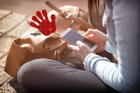 Kobieta z telefonem w dłoniach, obok brązowa torba i czerwona dłoń z logo heyah.