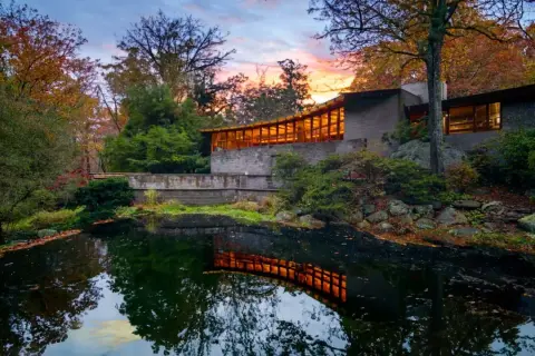 The last Wright house, a modern masterpiece, reflects warmly in a tranquil pond amidst autumn foliage.