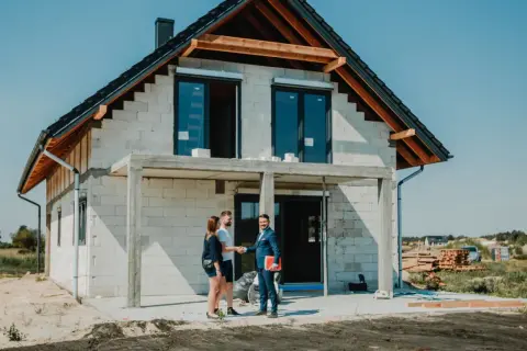 Para młoda uśmiecha się do doradcy, który wręcza im dokumenty. W tle widać dom w budowie, symbolizujący ich marzenie o własnym lokum, które zrealizują dzięki kredytowi na budowę domu.