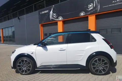 Biały Alpine A290 z niebieskimi akcentami stoi przed budynkiem z pomarańczowymi drzwiami garażowymi.