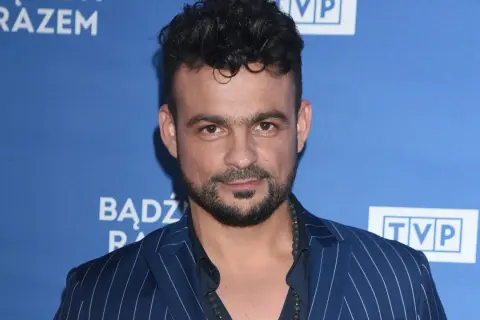 Robert El Gendy w granatowej marynarce w paski, z charakterystyczną fryzurą i zarostem, pozuje na tle niebieskiego banera z logo TVP.