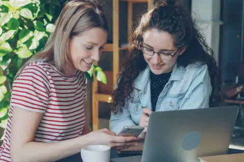 Dwie młode kobiety z proaktywną postawą pracują razem przy laptopie i telefonie, otoczone zielenią.