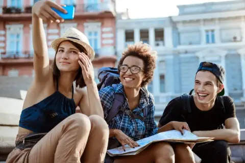 Tres amigos sonríen mientras se toman una selfie en la calle, planeando cómo conocer gente para viajar y explorar la ciudad.