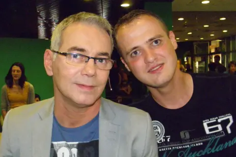 Dwóch mężczyzn pozuje do zdjęcia. Starszy, w okularach i jasnej marynarce, zastanawia się, czy prowadzący naprawdę zna wszystkie piosenki, czy korzysta z promptera.