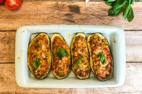 Quatre moitiés d'aubergines farcies, gratinées et garnies de basilic, dans un plat blanc sur fond de bois.