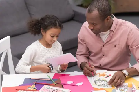 A father and daughter are crafting Mother's Day cards. The toddler cutting paper with scissors is focused on her task.