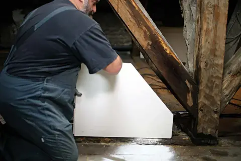 Un ouvrier installe un cloison carreau de plâtre blanc près d'une poutre en bois ancienne.