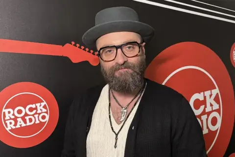 Mężczyzna w kapeluszu i okularach, z brodą, na tle logo Rock Radia. Widać napis "perfect wszystko ma swój czas tekst".