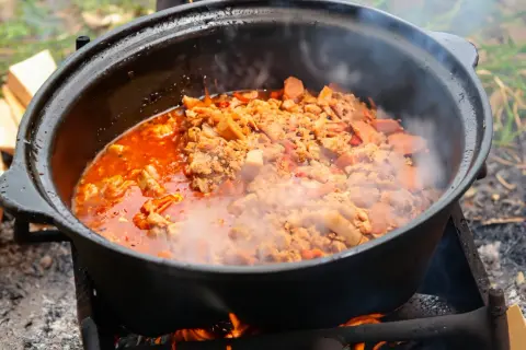 Herzhafte Eintopf-Rezepte im Kessel über offenem Feuer. Dampf steigt auf, während Fleisch und Gemüse in der roten Sauce köcheln.