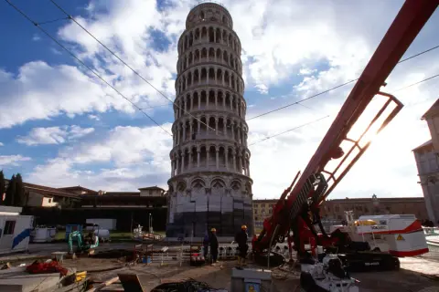 La Torre di Pisa prima e dopo i lavori di consolidamento: un cantiere con macchinari e operai accanto alla torre pendente.