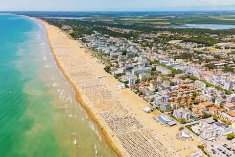 Słoneczna bibione plaża z tysiącami leżaków i parasoli, turkusowe morze i miasto w tle.