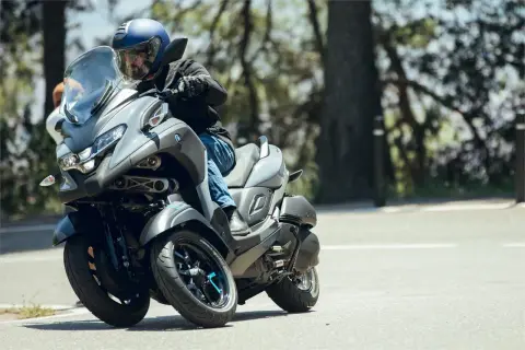 Hombre en casco azul conduce un triciclo Yamaha Tricity 500 por una carretera arbolada, inclinándose en una curva.
