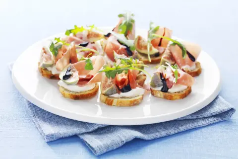 Köstliche italienische Crostini mit Prosciutto, Feigen, Mozzarella und Rucola auf einem weißen Teller.