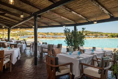 Tavoli apparecchiati sotto un tetto di paglia, con vista sulla piscina e sulla spiaggia del **hotel Cala di Volpe**.