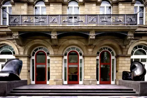 Vor dem Museum Wuppertal stehen zwei abstrakte Skulpturen. Die Fassade des Gebäudes ist aus hellem Stein, mit roten Türen und verzierten Balkonen.