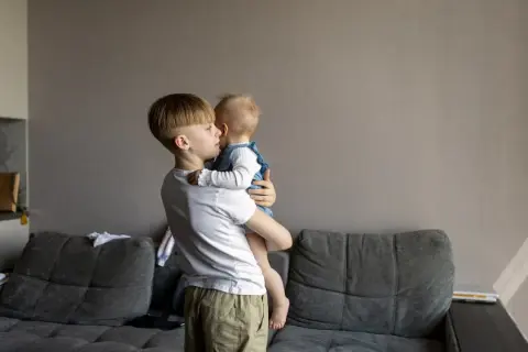 Starszy brat trzyma na rękach niemowlę. Zastanawiamy się, od jakiego wieku dziecko może zostać samo w domu, ale ta troska jest widoczna w jego spojrzeniu.