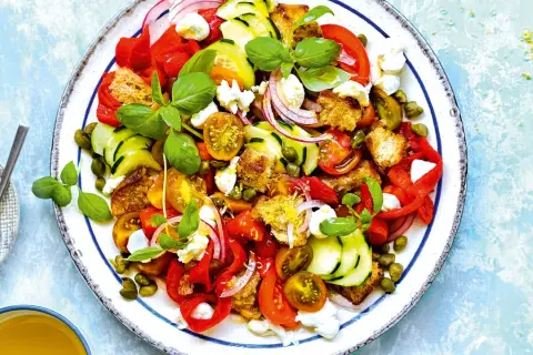 Ensalada de burrata vibrante con tomates, pepino, pimientos rojos, pan tostado y alcaparras, adornada con albahaca fresca.