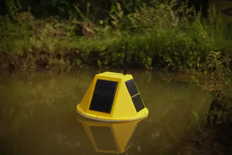 Boya amarilla con paneles solares y antena flotando en agua turbia, parte de un sistema de control de nivel de agua.
