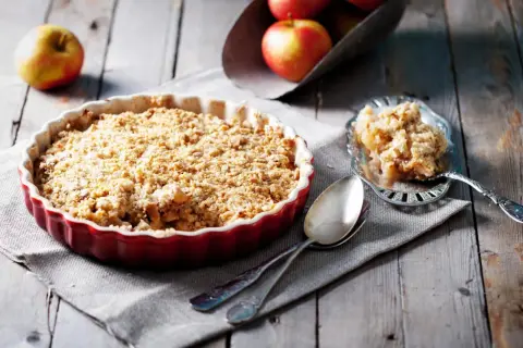 Un délicieux crumble aux pommes tout juste sorti du four, servi dans un plat rouge, avec une portion sur une cuillère ornée.