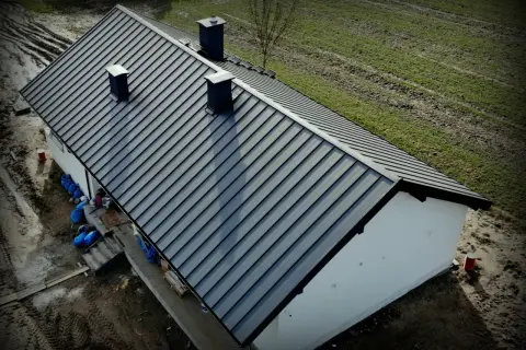 Nowy, ciemnoszary dach z blachy na domu jednorodzinnym. Widać kominy i otoczenie budowy. Dobry koszt dachu to podstawa.