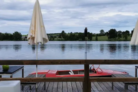 Gemütliches Restaurant am See mit rotem Tretboot und Sonnenschirmen. Die Uferlandschaft ist grün und hügelig.