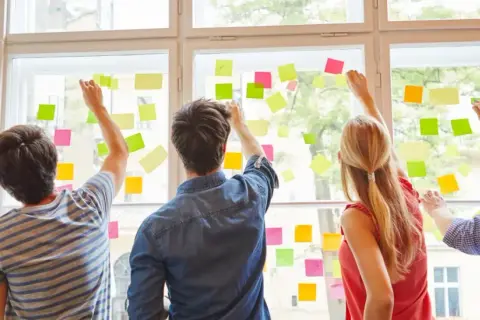 Fünf Personen planen an einem Fenster mit bunten Haftnotizen. Sie arbeiten an ihrer kaufmännischen Ausbildung nebenberuflich.