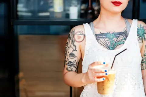 Junge Frau mit vielen Tattoos, darunter ein Vogel und ein Auge. Sie hält einen Eiskaffee. Ein Tattoo ab 16, das ihre Persönlichkeit unterstreicht.