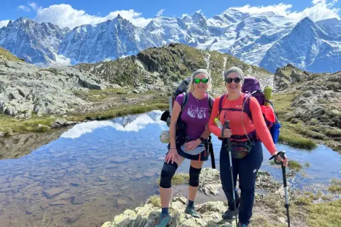 Dwie kobiety cieszą się trekkingiem wokół Mont Blanc. Mają plecaki i kije trekkingowe, a w tle widać ośnieżone szczyty.