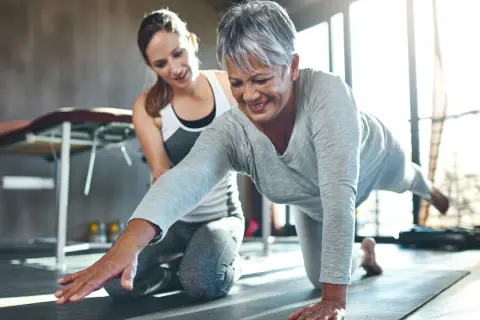 Une femme âgée sourit en faisant un exercice de renforcement avec l'aide d'une coach. Le sport est essentiel pour la rééducation après un cancer du sein.