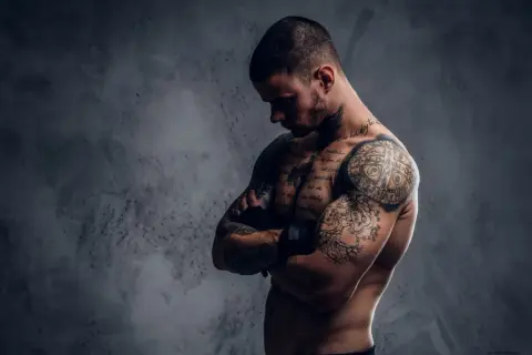 Ein muskulöser Mann mit vielen Tattoos, der seine Arme verschränkt. Sein Körperbau zeigt, dass er viel für Sport und Training tut.