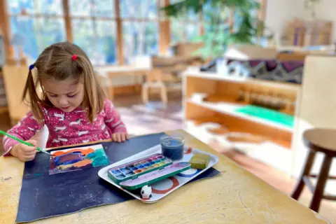A young girl with pigtails paints at a table, exploring her creativity with watercolors. This scene captures the essence of Montessori nature-inspired learning.