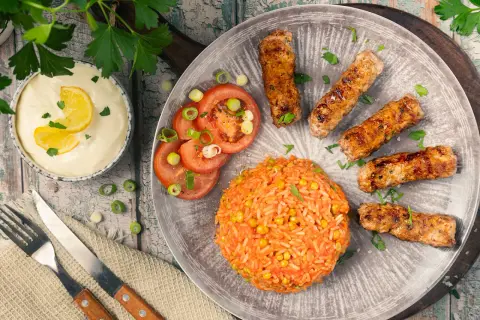 Saftige cevapcici im backofen, serviert mit Reis, Tomaten und einer cremigen Sauce.