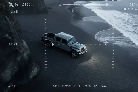 Un camion du futur gris, vu du ciel, roule sur une plage de sable noir. Les vagues de l'océan s'écrasent sur le rivage.